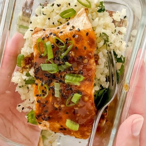 Two hands holding a glass meal prep container of salmon on top of cauliflower rice garnished with green onions and sesame seeds.