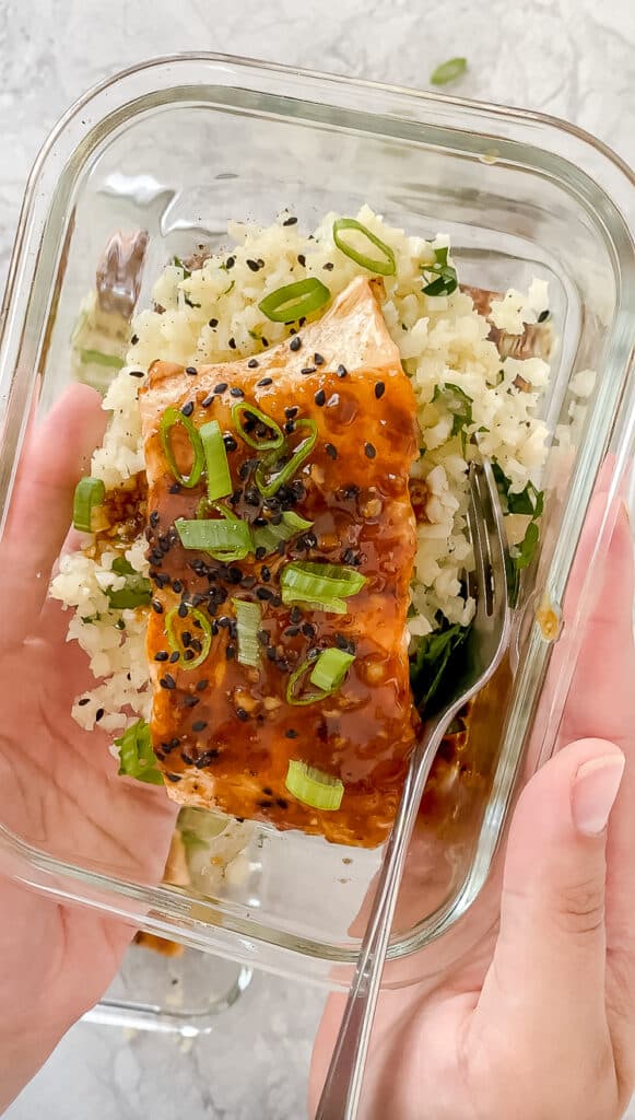 Two hands holding a glass meal prep container of salmon on top of cauliflower rice garnished with green onions and sesame seeds.