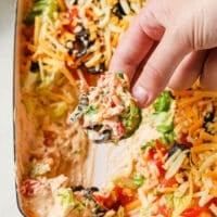 Hand holding a cucumber slice with the taco dip over the remaining dish atop a marble counter.