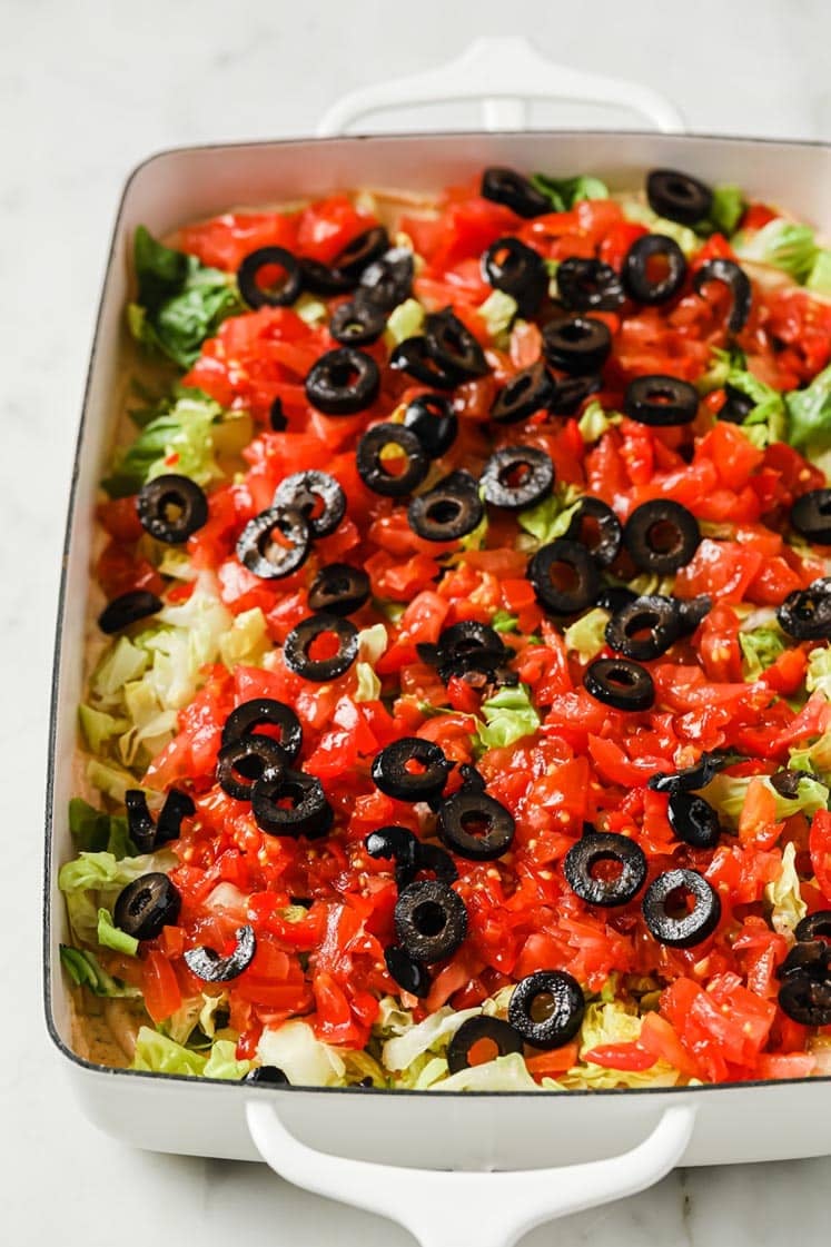 A white rectangular dish filled with layered salad ingredients, including chopped lettuce, diced tomatoes, and sliced black olives. The vegetables are colorful and evenly spread across the top.