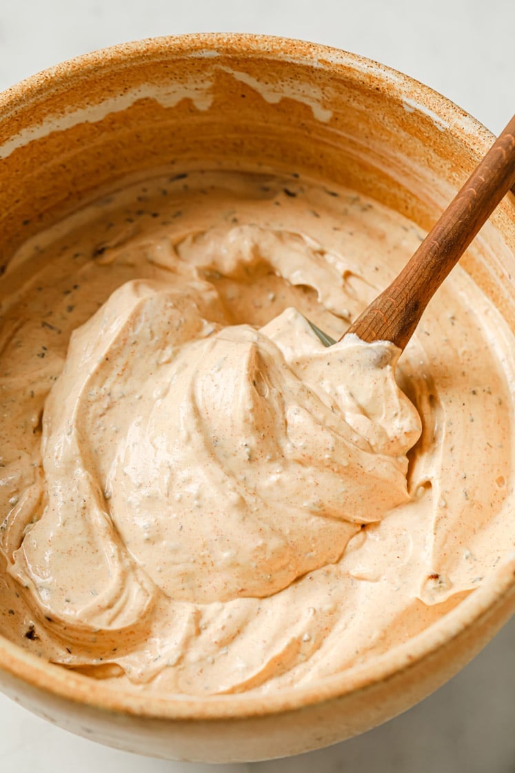 A wooden spoon stirring a creamy, light orange sauce with herbs in a beige ceramic bowl.