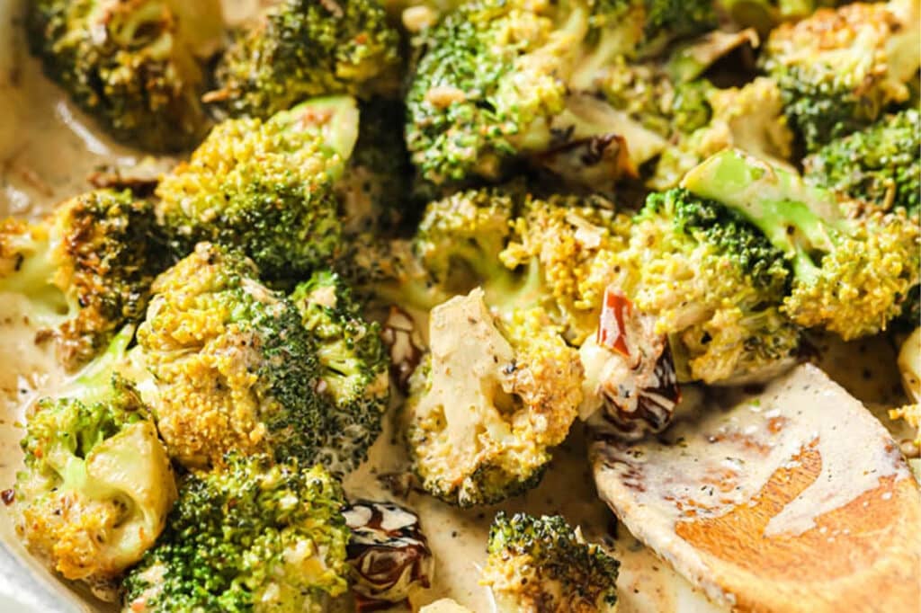 Close-up of Creamy Italian Broccoli With Sun-Dried Tomatoes garnished with black pepper in a large pan with a wooden spatula.