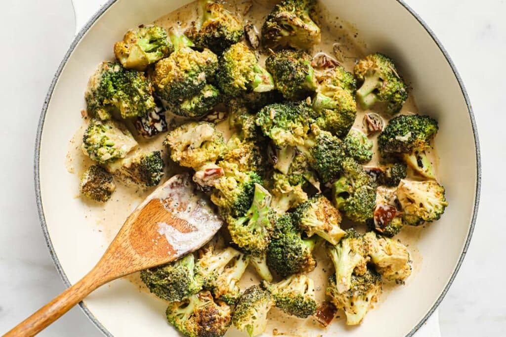 Creamy Italian Broccoli With Sun-Dried Tomatoes garnished with black pepper in a large pan with a wooden spatula.