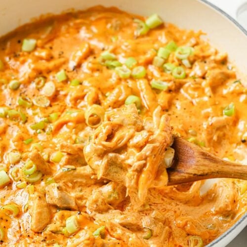 Green Chile Chicken With Leftover Shredded Chicken Thighs garnished with chopped green onions and black pepper in a cast iron skillet pan with a wooden spatula for serving atop a marble counter.