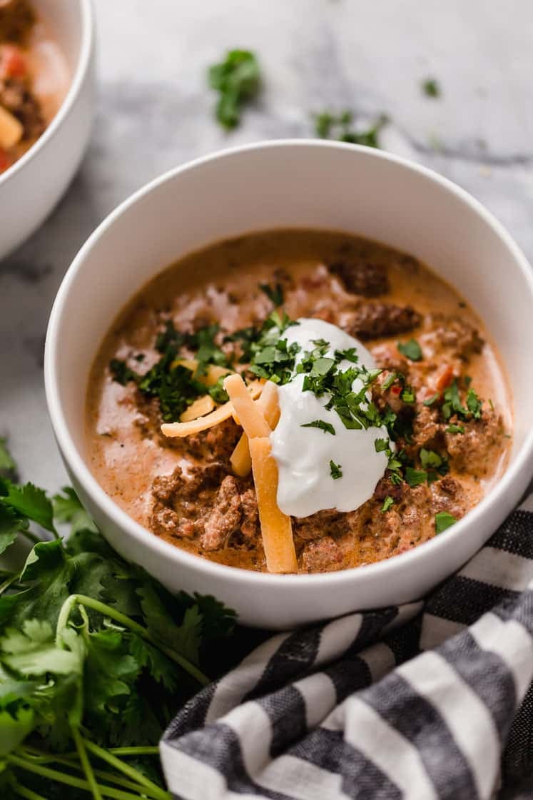 Crock Pot Taco Soup garnished with a dollop of sour cream, grated cheddar cheese, and freshly chopped parsley served in bowls beside sprigs of fresh parsley and a striped table napkin atop a marble counter.
