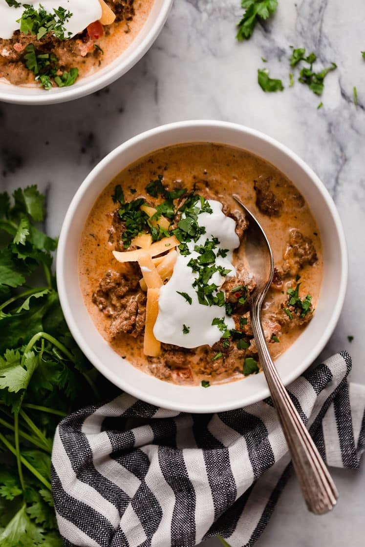 Crock Pot Taco Soup garnished with a dollop of sour cream, grated cheddar cheese, and freshly chopped parsley served in bowls with a spoon beside sprigs of fresh parsley and a striped table napkin atop a marble counter.