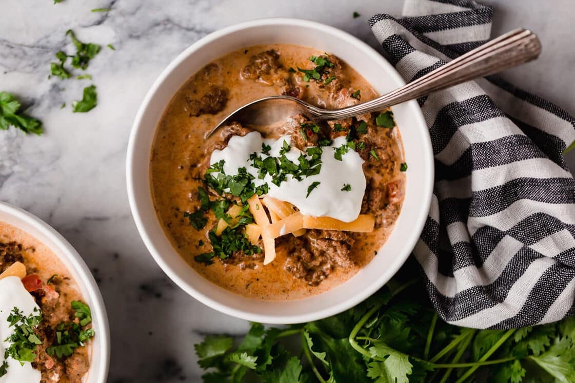 Crock Pot Taco Soup garnished with a dollop of sour cream, grated cheddar cheese, and freshly chopped parsley served in bowls with a spoon beside sprigs of fresh parsley and a striped table napkin atop a marble counter.