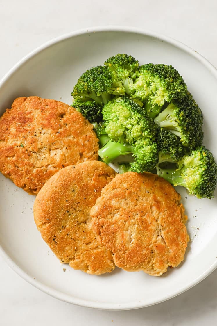 Canned Chicken Patties and steamed broccoli florets garnished with freshly cracked black pepper on a plate atop a marble counter.