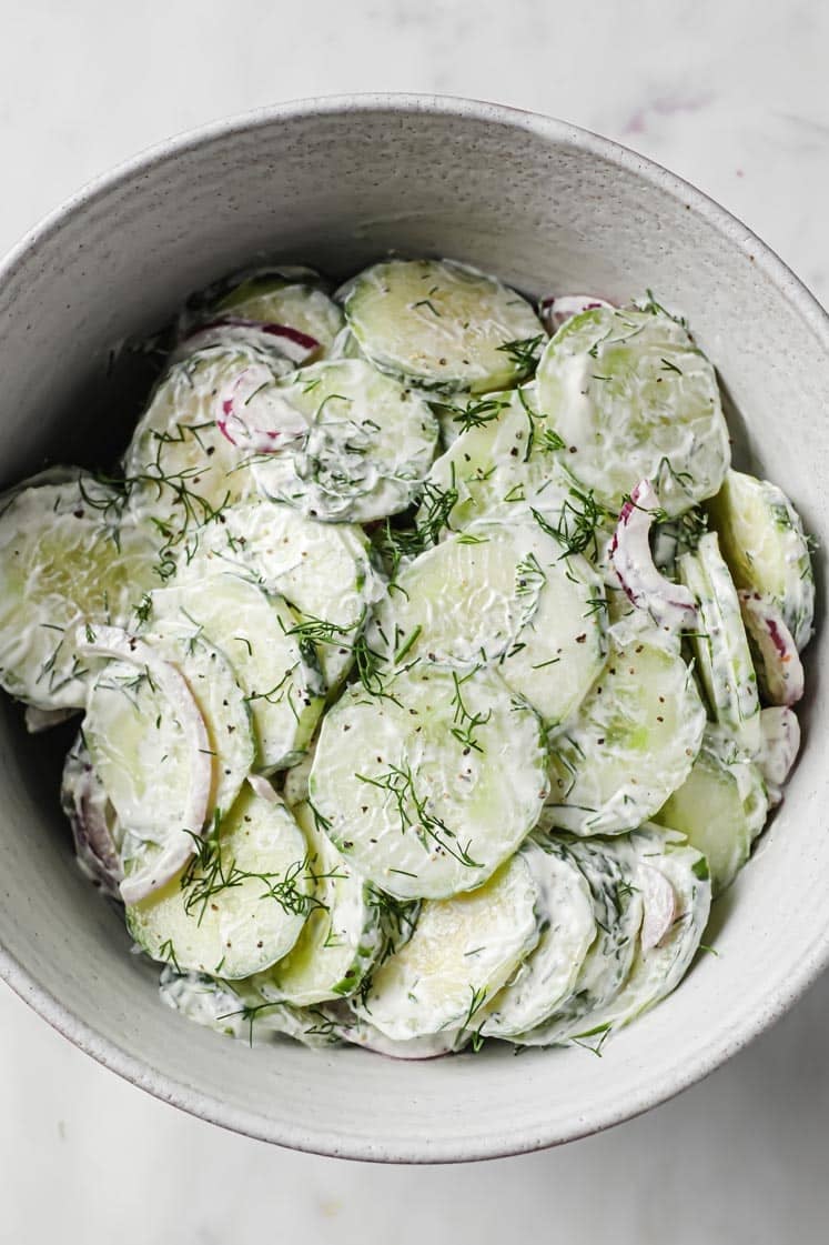 Creamy Dill Cucumber Onion Salad (With Sour Cream And Mayo) in a ceramic bowl atop a marble counter.