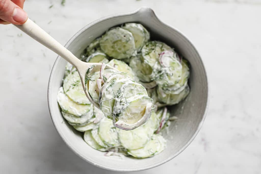 Close-up of a portion of Creamy Dill Cucumber Onion Salad (With Sour Cream And Mayo) on a spoon over a bowl of the same dish atop a marble counter.