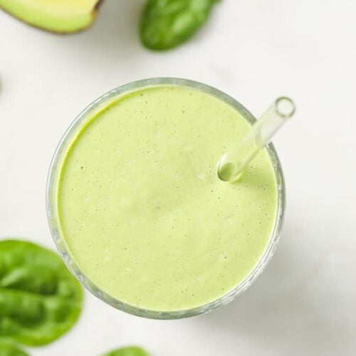 Overhead shot of the Keto Green Smoothie on a glass with clear straw, along with half of an avocado and scattered spinach leaves on the marble countertop.