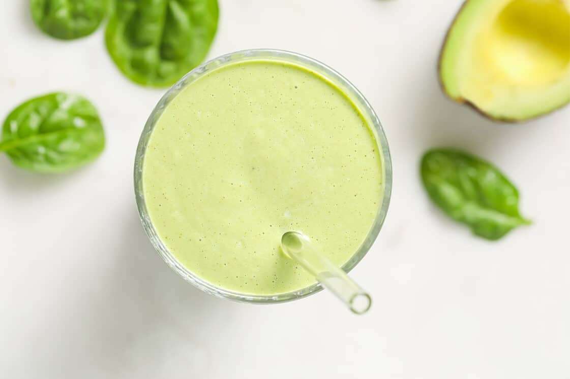 Overhead shot of the green smoothie in a glass with a clear straw, next to half an avocado and scattered spinach leaves on a marble countertop.