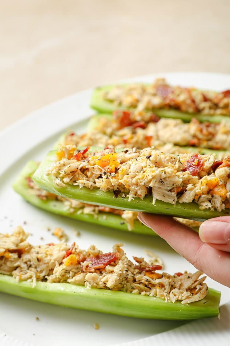 A hand lifting one bacon ranch chicken salad cucumber boat off a white plate holding the rest.