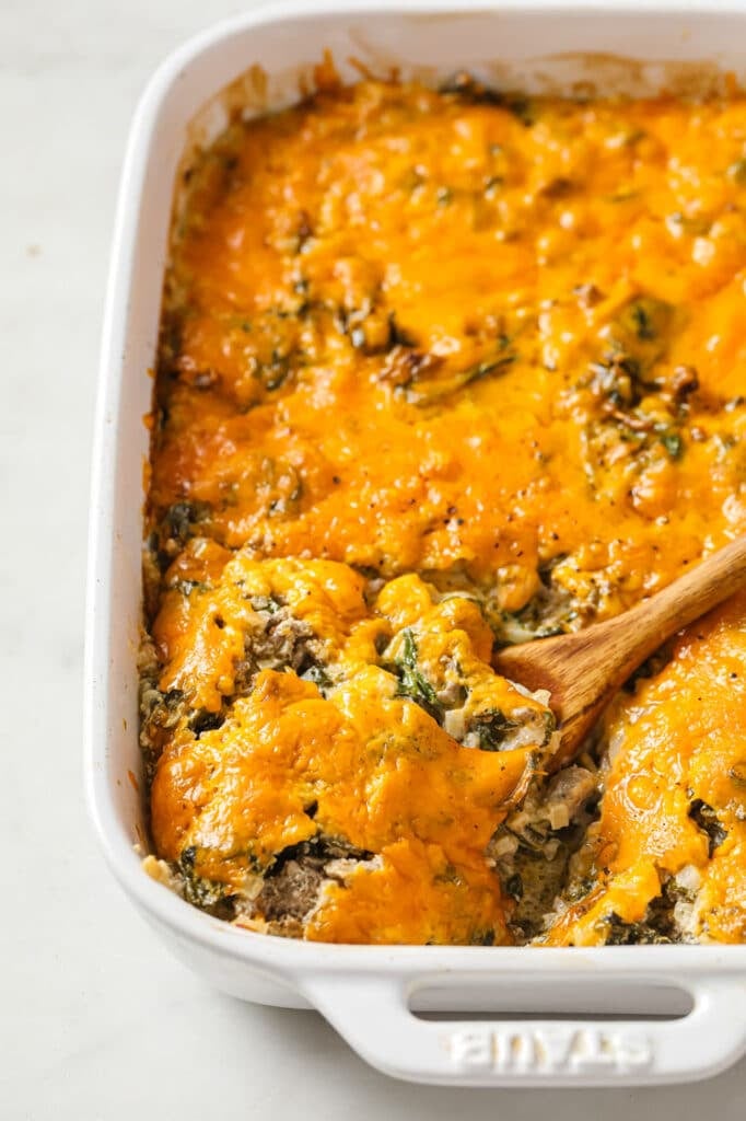 Cheesy beef casserole in a baking dish with a wooden serving spoon atop a marble countertop.