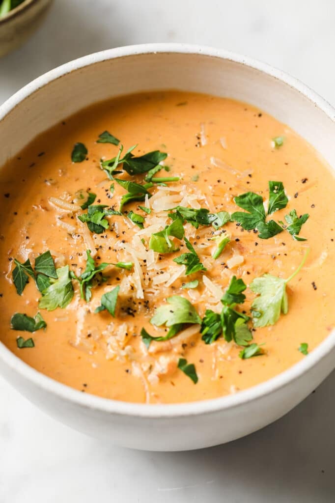 A serving of garnished Creamy Italian Sausage Soup in a white bowl beside a small bowl with chopped parsley on a marble countertop.