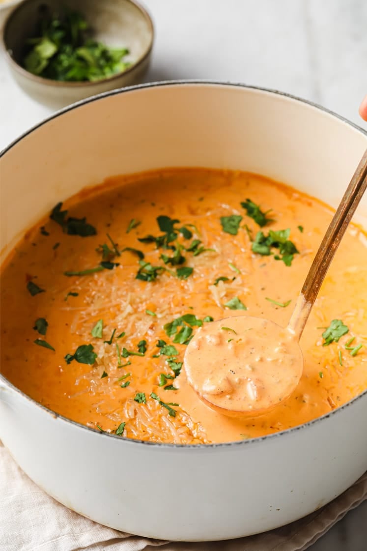 Garnished Creamy Italian Sausage Soup in a white pot with a ladle scooping a portion, resting on a white kitchen towel beside a small bowl with chopped parsley on a marble countertop.