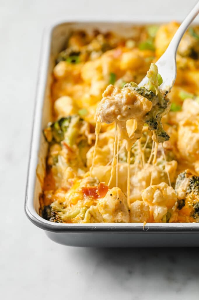 Loaded broccoli cauliflower casserole in a baking dish with a portion on a fork with melty cheese hanging down.
