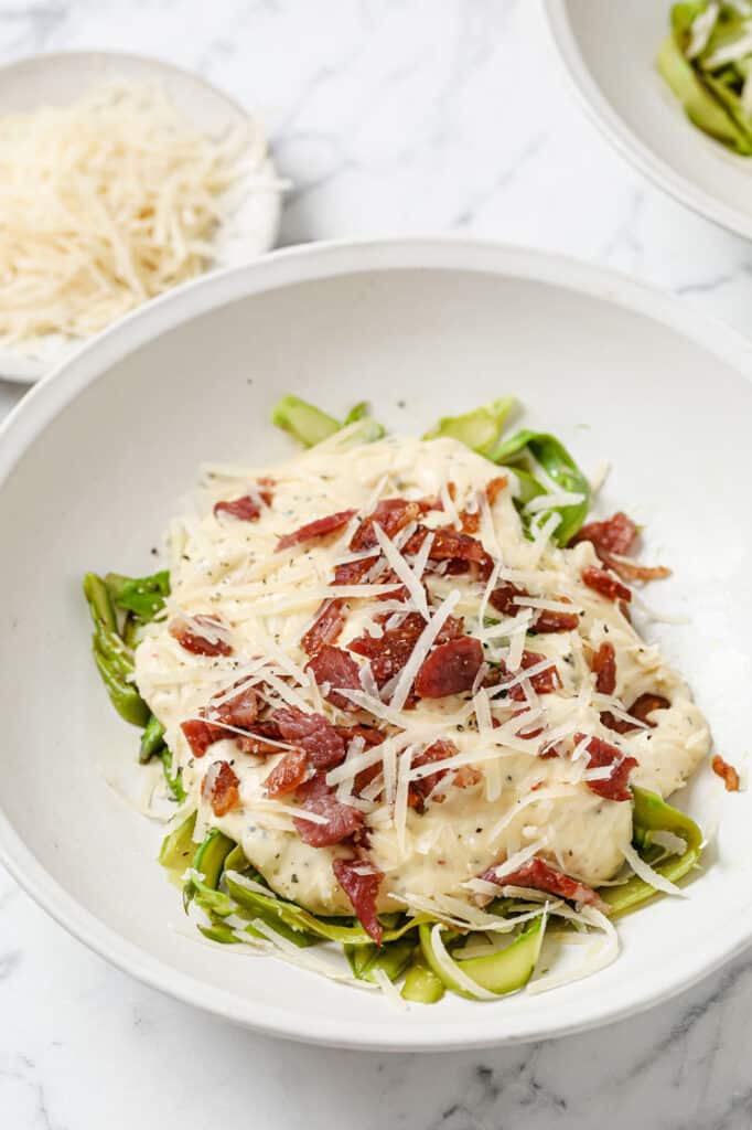 Asparagus noodles with alfredo sauce and bacon in a bowl and garnished with cheese.