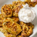 A plate of baked zucchini bites served with a dipping sauce. One of the zucchini bites is being dipped in the sauce.