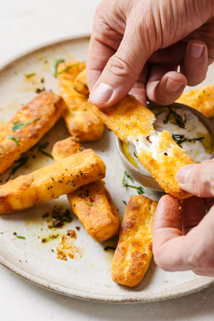 A person holding one halloumi fry over a plate of the same dish.