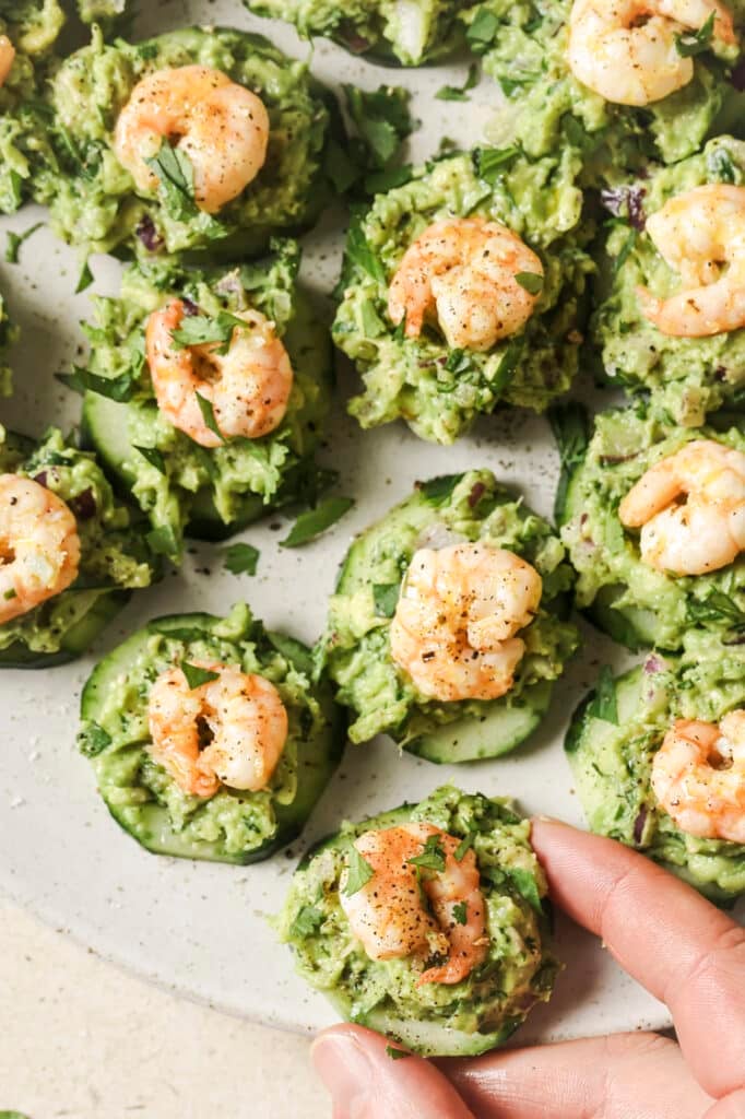 A plate of garnished cold shrimp and cucumber appetizer with a person holding a piece.