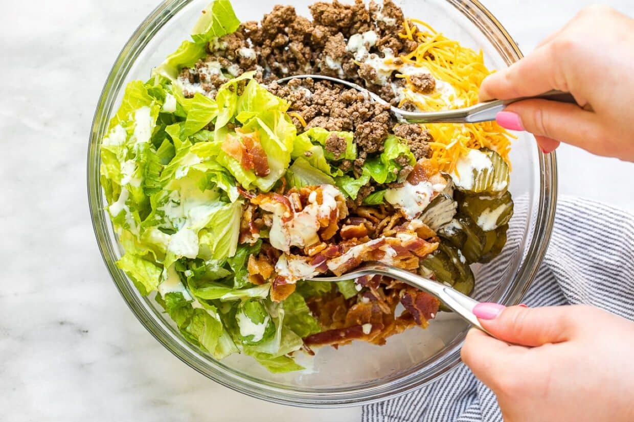 A person holding serving spoons in a bowl of Big Mac salad.