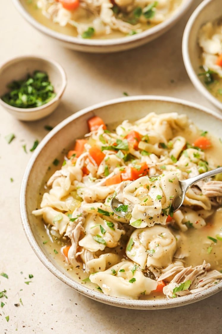 Chicken tortellini soup in bowls, with one being scooped by a spoon.