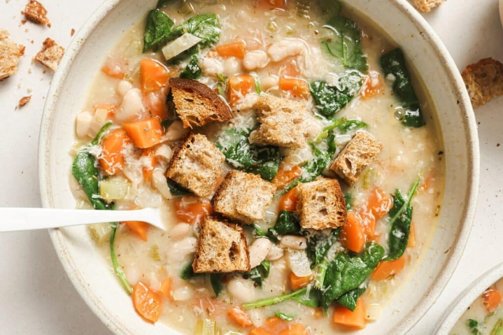 A bowl of vegetable soup with white beans, carrots, spinach, and croutons.
