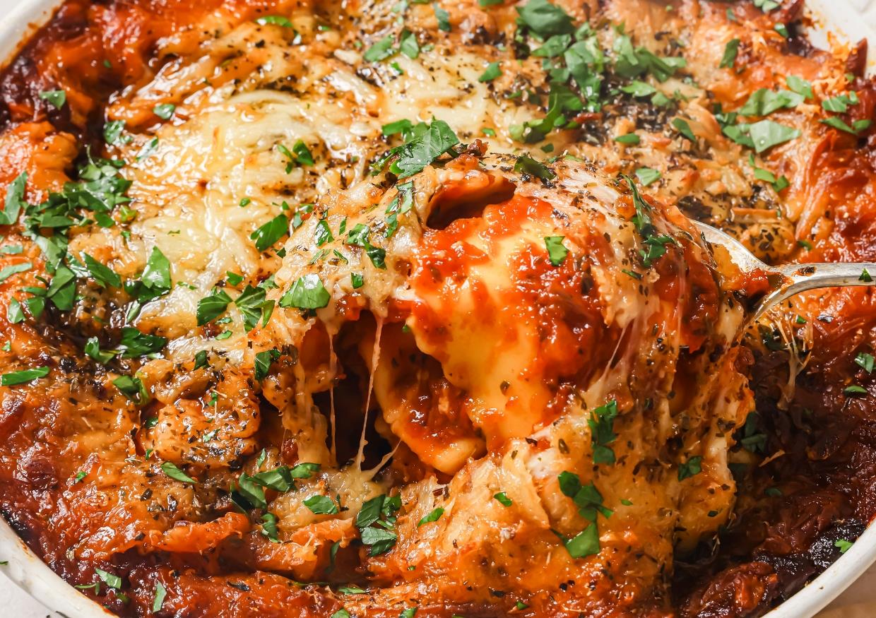 A close-up of a cheesy baked ravioli garnished with herbs, with a fork lifting a portion to show the melted cheese.