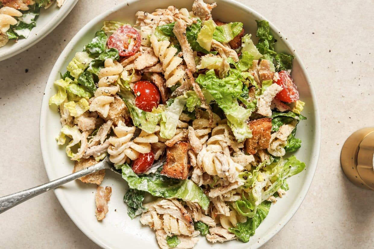 A fresh Caesar salad with chicken, croutons, tomatoes, and dressing, served in a large white bowl with a fork.