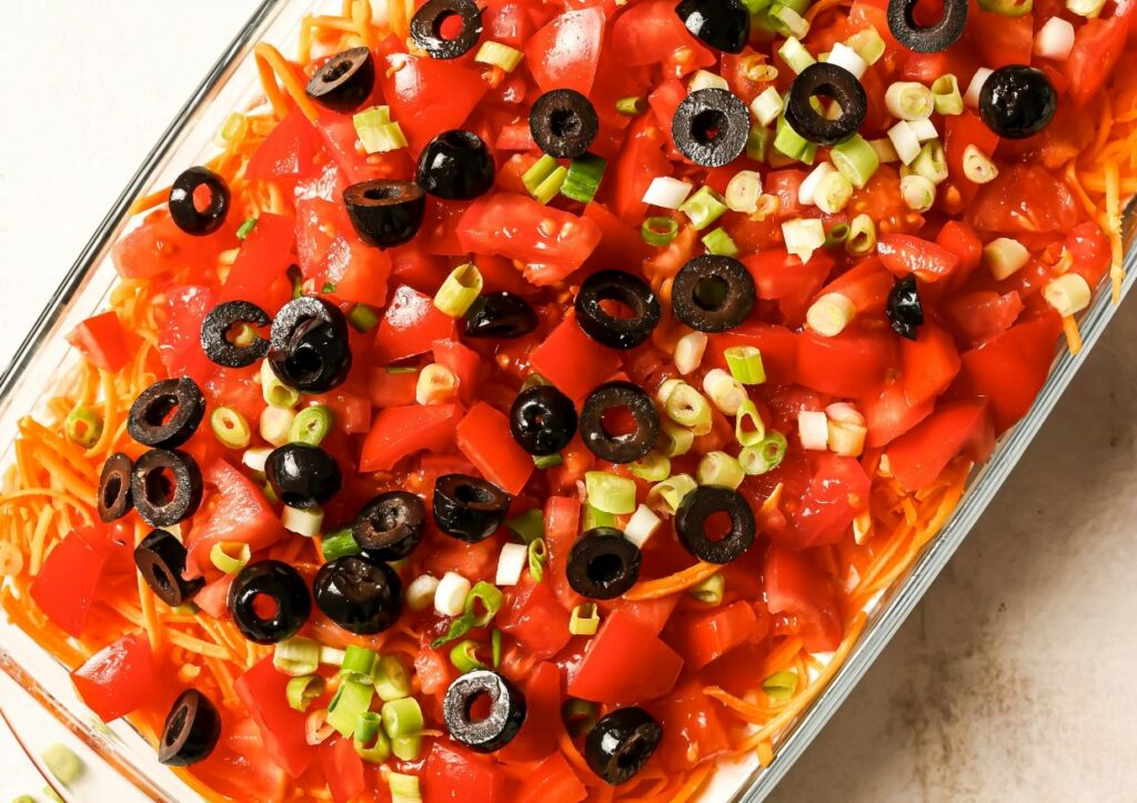 A rectangular dish filled with a layered dip, featuring shredded cheese, chopped tomatoes, black olive slices, and green onions.