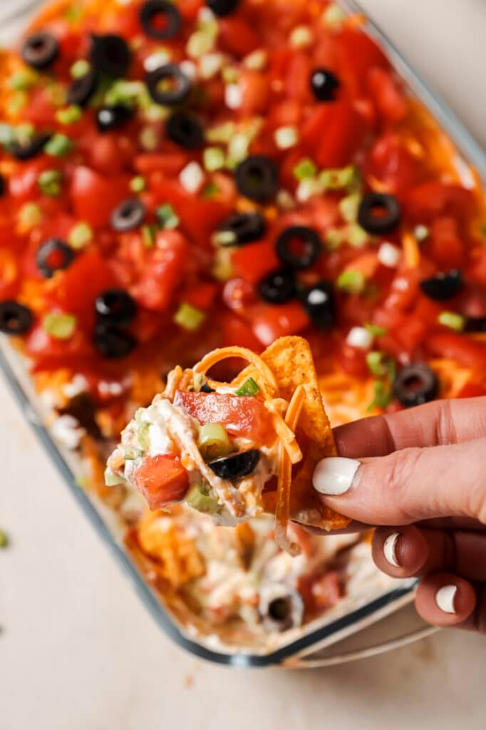 A hand holding a tortilla chip with 7-layer dip over the baking dish with the rest of the dip.