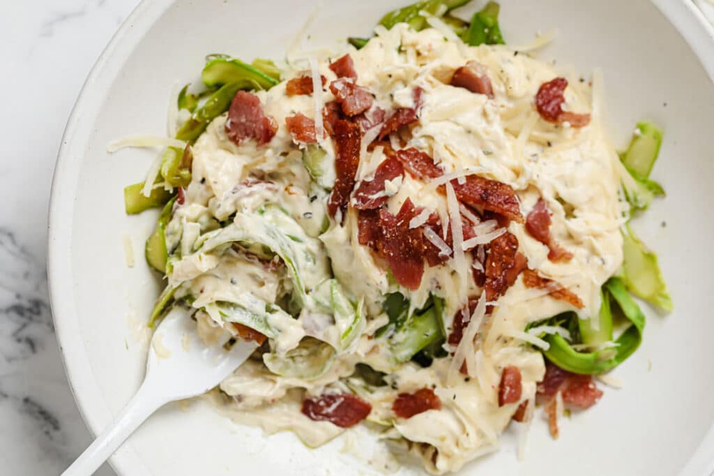 Asparagus noodles with Alfredo sauce and bacon on a plate with a fork.