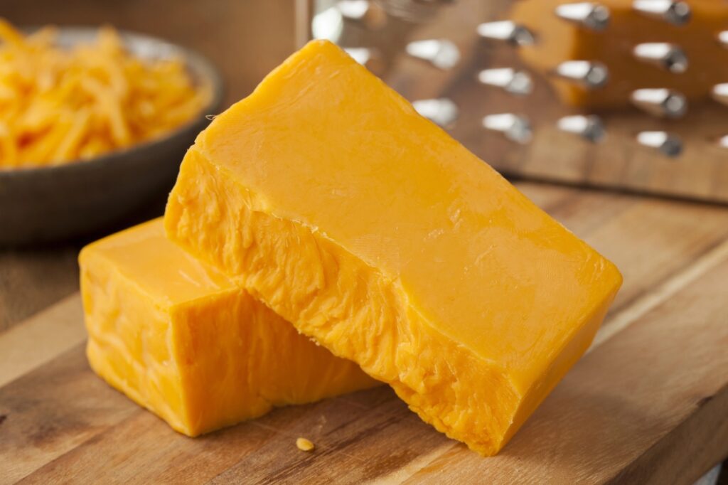 Two blocks of cheddar cheese near a bowl of shredded cheese and a cheese grater.
