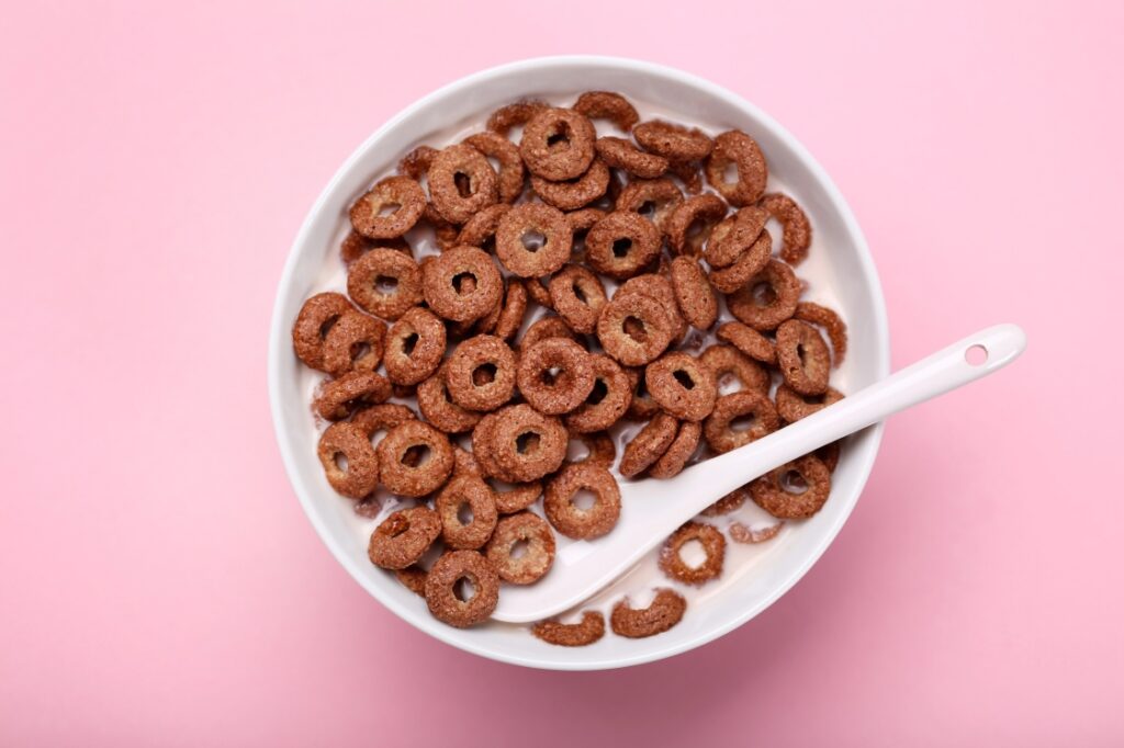 A bowl of cereal and milk with a spoon on a pink background.
