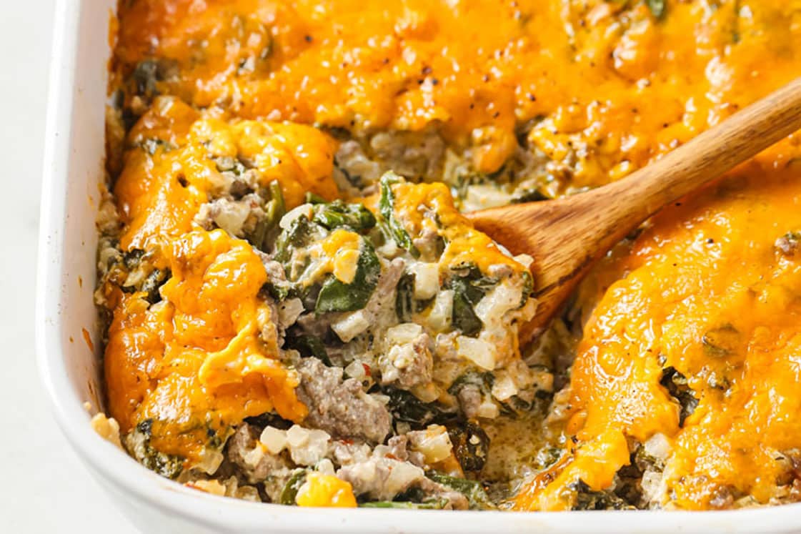 Cheesy Beef Casserole With Cauliflower Rice And Spinach on a baking dish with a wooden serving spoon atop a marble countertop.