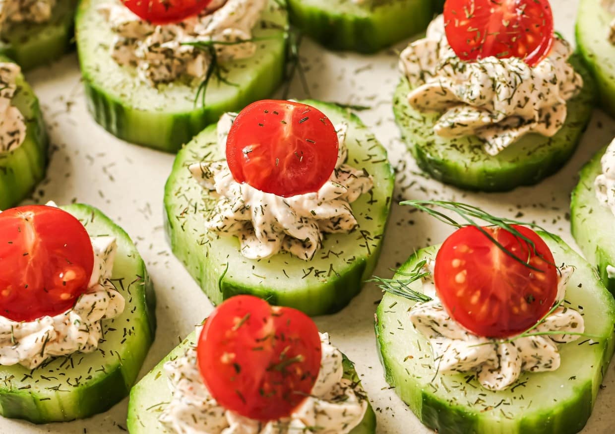 Sliced cucumbers topped with dollops of herb cream cheese, cherry tomato halves, and dill sprigs arranged on a beige surface.