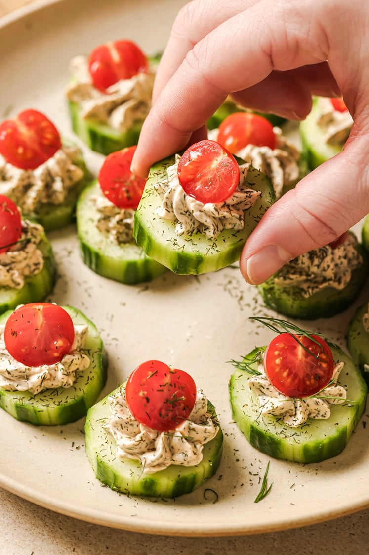 Garnished cucumber tomato bites on a plate, one held by a hand.