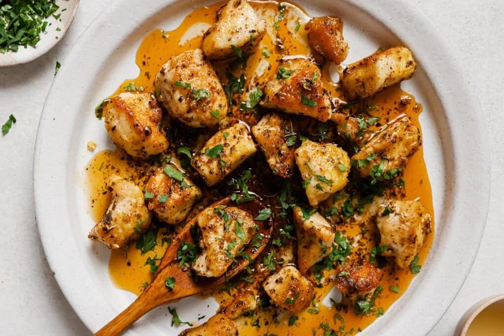 Garnished garlic butter chicken bites on a plate with a piece on a wooden spoon.