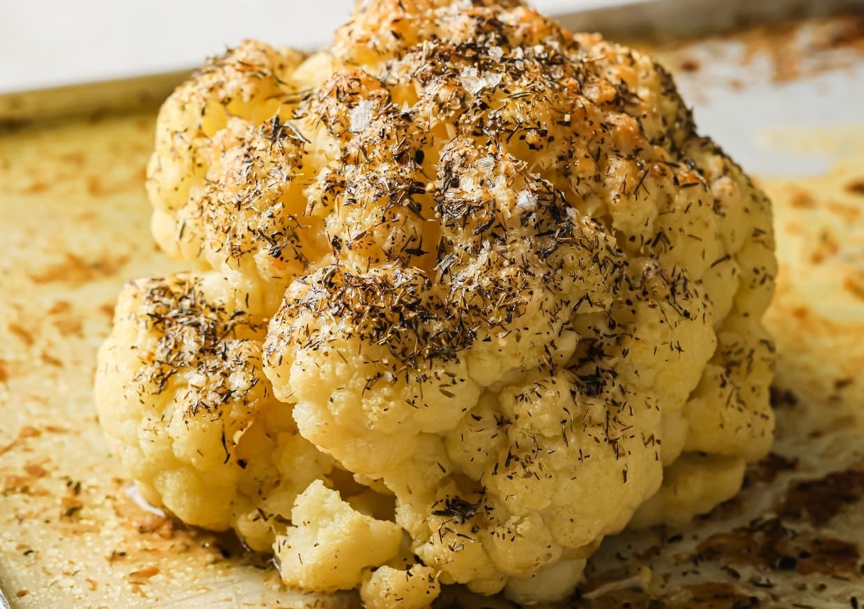 A roasted whole cauliflower head topped with herbs and seasoning on a baking tray.