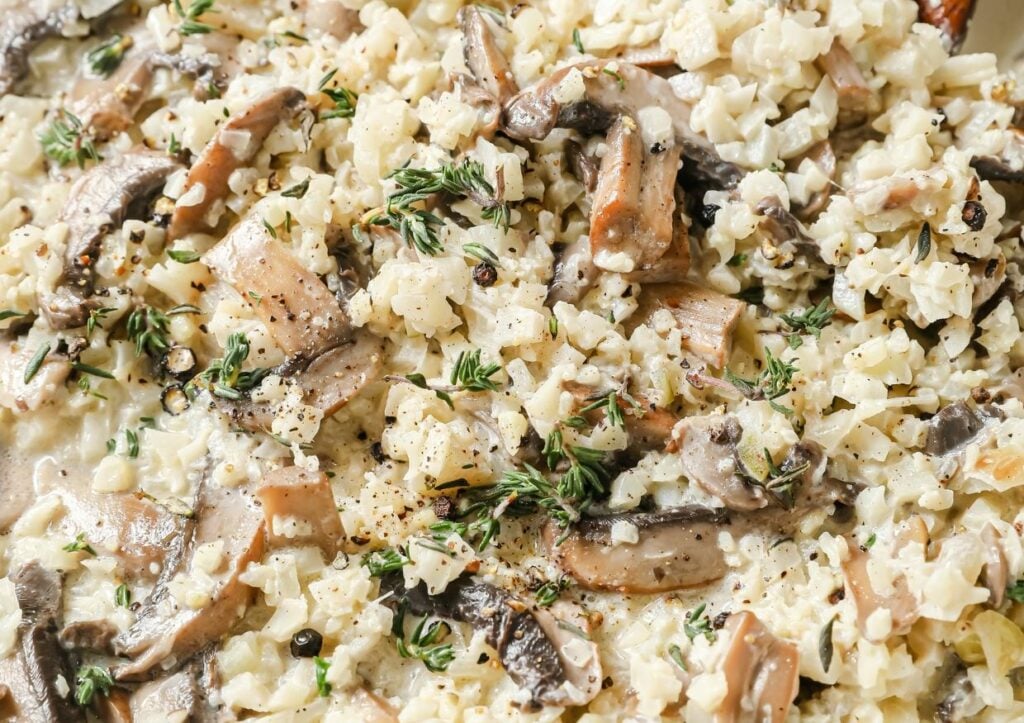 A close-up of a creamy cauliflower risotto with mushroom pieces, garnished with fresh thyme and black pepper.