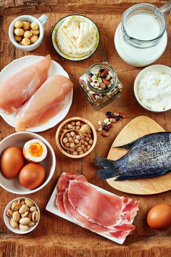 Assorted protein sources like chickpeas, chicken breasts, eggs, cheese, nuts, fish, milk, and more on a wooden board.