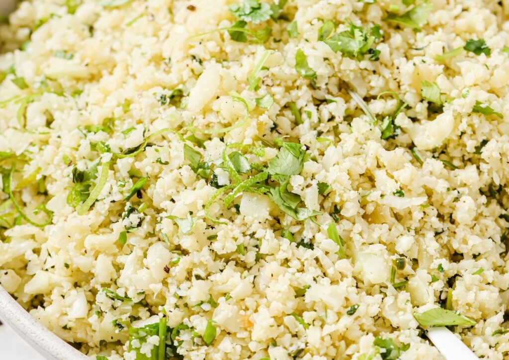 Close-up of a dish consisting of cauliflower rice mixed with herbs and seasonings. The finely grated cauliflower resembles rice grains, and green herbs are sprinkled throughout the dish.