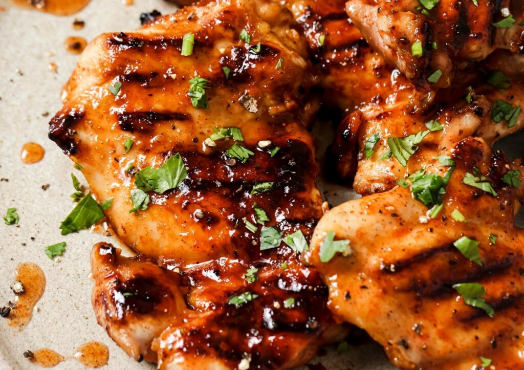 Close-up of grilled chicken pieces garnished with chopped herbs and drizzled with a glossy sauce on a light-colored plate.