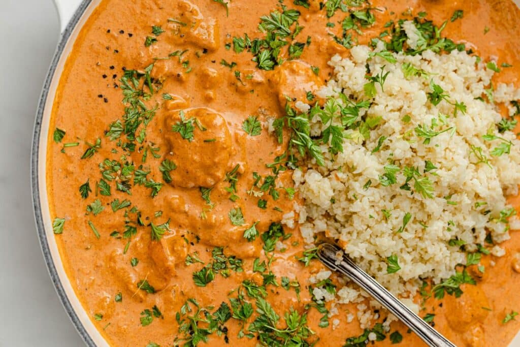 A plate of orange-colored butter chicken garnished with chopped herbs, next to a serving of cauliflower rice, with a spoon partially visible.