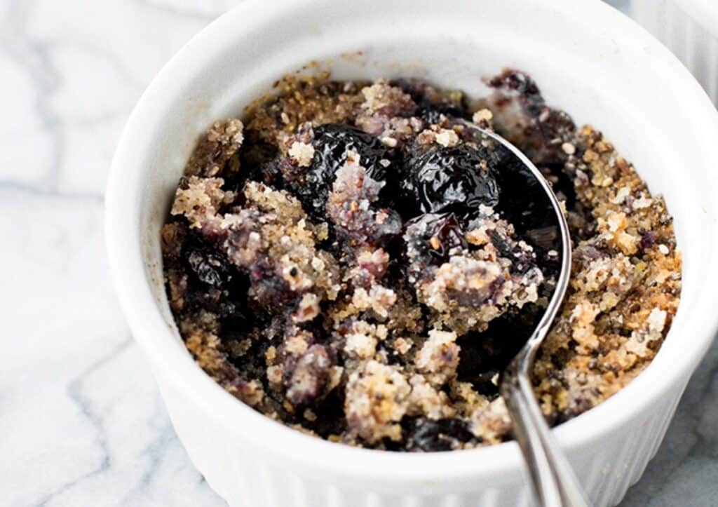 A close-up of a small white ramekin containing a blueberry crumble dessert with a metal spoon inside.