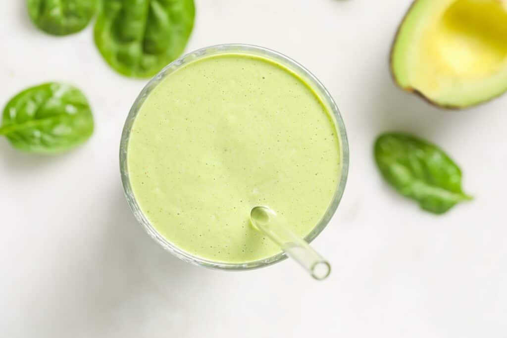 Top-down view of a green smoothie in a glass with a straw, surrounded by spinach leaves and half an avocado on a light surface.