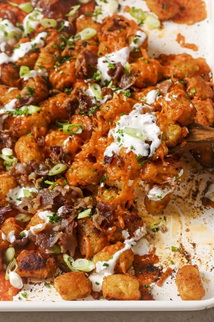 Loaded tater tots being scooped by a wooden spoon on a sheet pan.