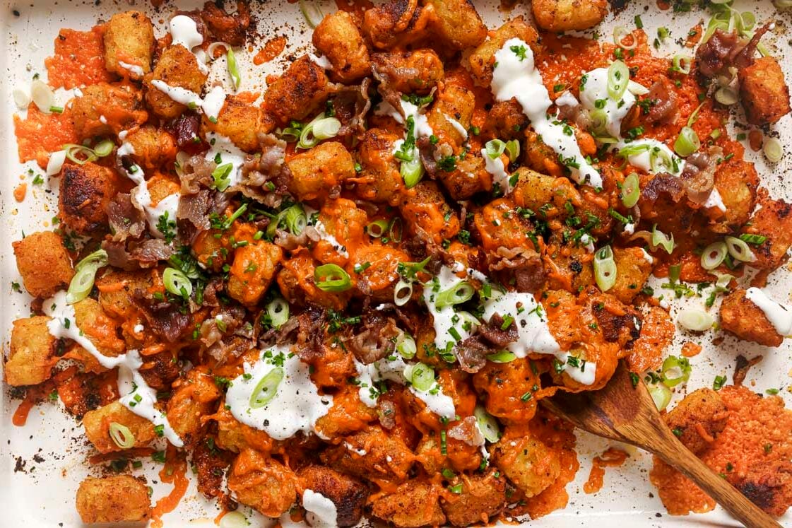 Loaded tater tots garnished with sour cream on a sheet pan with a wooden spoon.