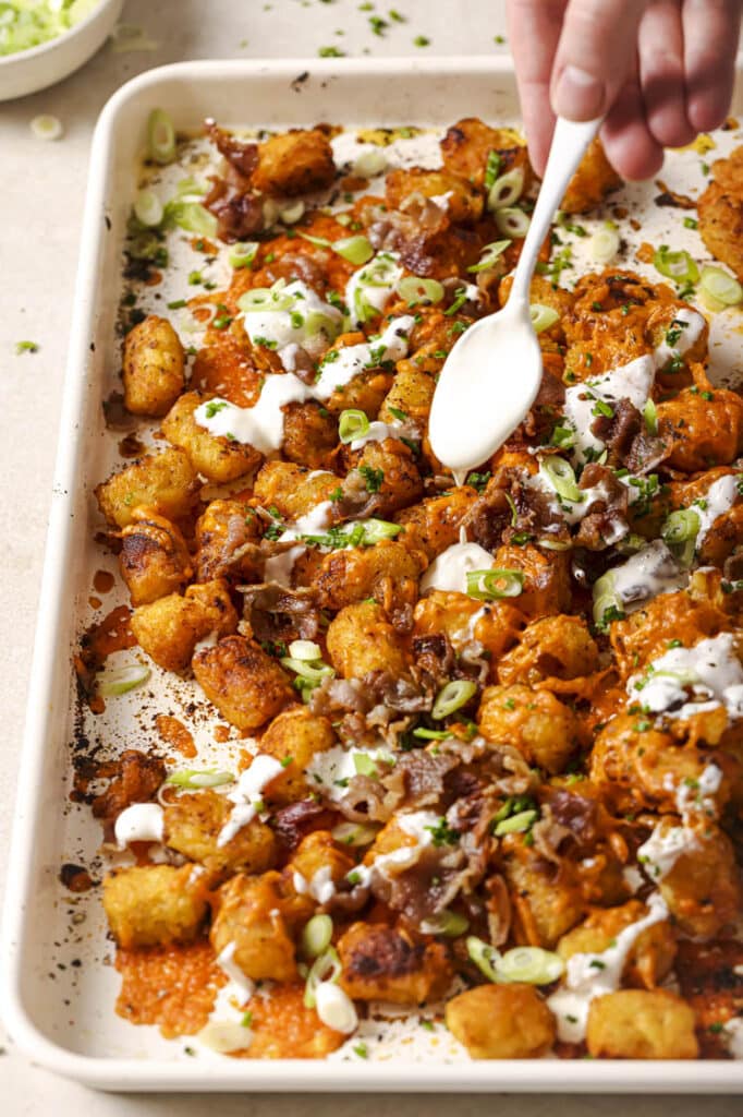 Loaded tater tots being garnished with sour cream on a sheet pan.